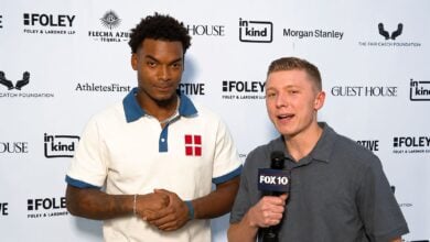 Arizona Cardinals cornerback Will Johnson stands with arms clasped next to FOX10 Phoenix reporter Blake Niemann during a one-on-one interview, serious expression on his face, against a sponsor backdrop in Phoenix.