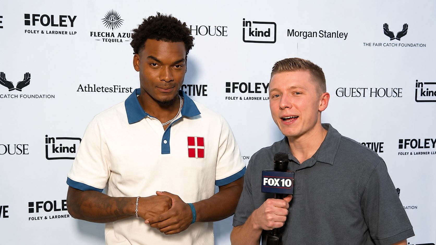 Arizona Cardinals cornerback Will Johnson stands with arms clasped next to FOX10 Phoenix reporter Blake Niemann during a one-on-one interview, serious expression on his face, against a sponsor backdrop in Phoenix.